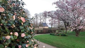 En grussti der fører hen til en bygning med mørkt tag, omgivet af græs og blomstrende træer.