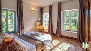 Et soveværelse med to senge, en fletstol, en lampe og store vinduer med gardiner.