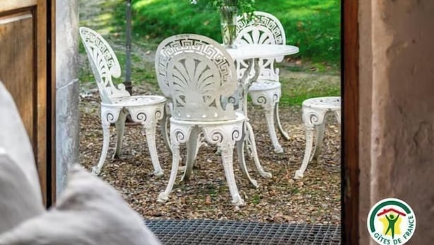 En døråbning der fører til et udendørs siddeområde med hvide metalstole og et bord, en hvid parasol og udsigt til en have.