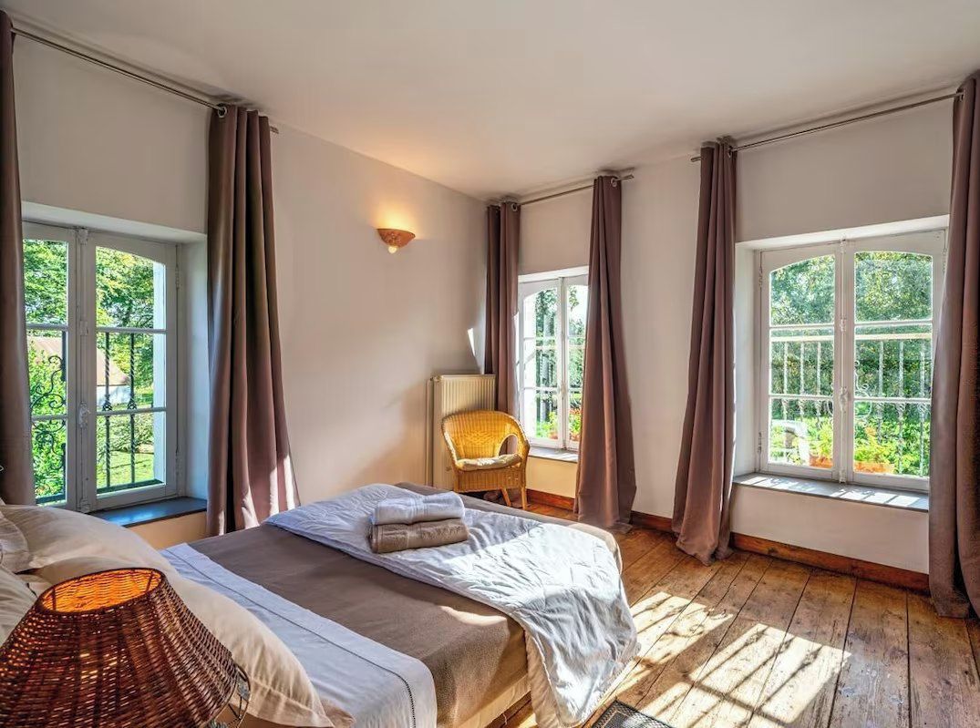 Un dormitorio con dos camas, una silla de mimbre, una lámpara y ventanas amplias con cortinas.