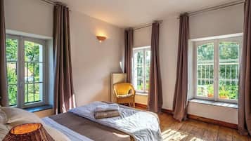 Un dormitorio con dos camas, una silla de mimbre, una lámpara y ventanas amplias con cortinas.