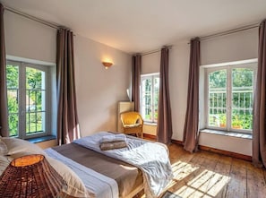 Een slaapkamer met twee bedden, een rieten stoel, een lamp en grote ramen met gordijnen.