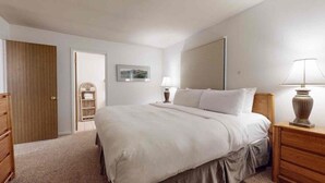 Une chambre avec un grand lit, une commode en bois, une table de chevet avec une lampe, une porte et une étagère fixée au mur où sont rangées des serviettes.