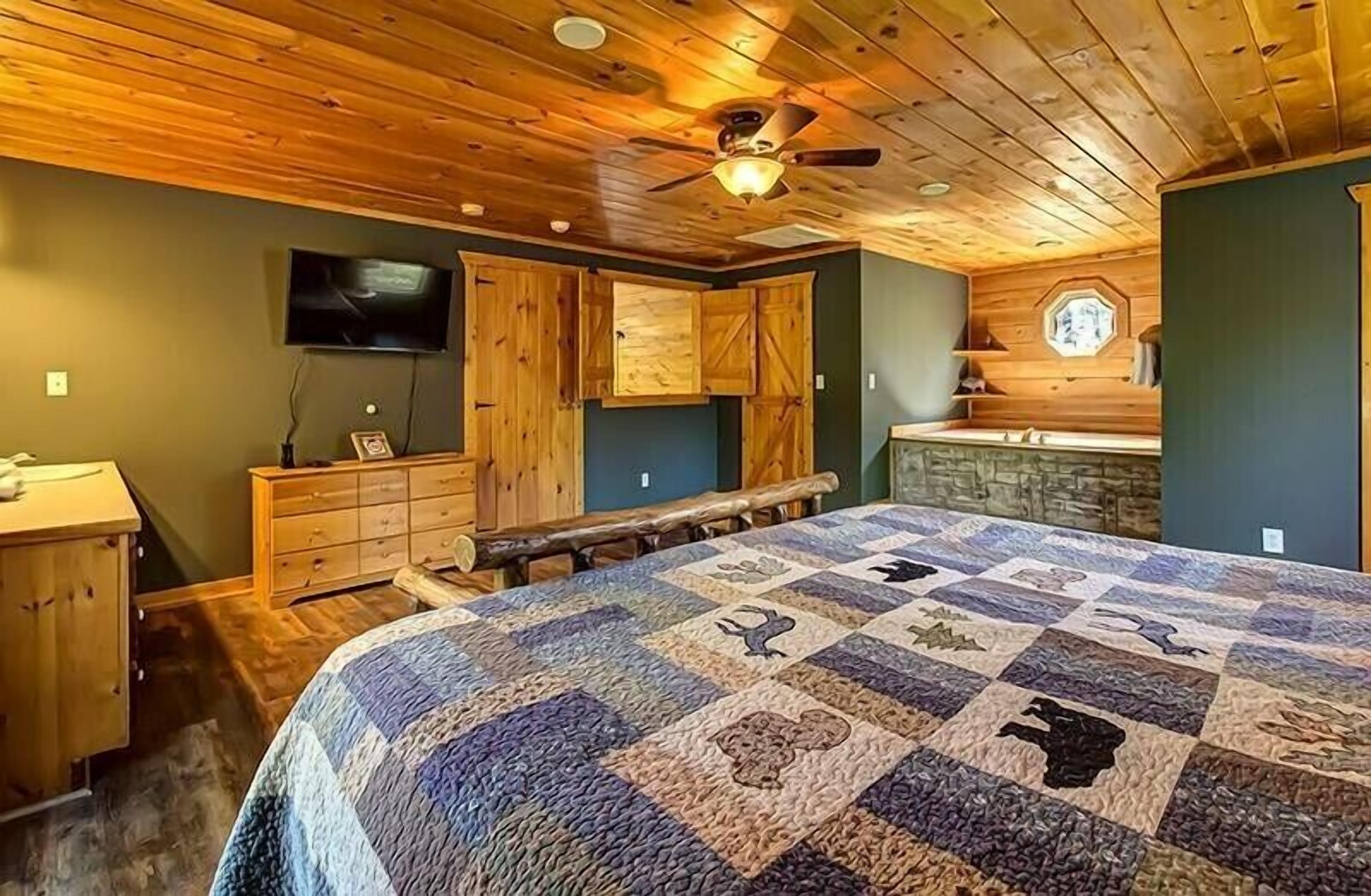 A cozy bedroom with a wooden ceiling, a large bed with a quilt, a wooden bench, and a TV mounted on the wall.