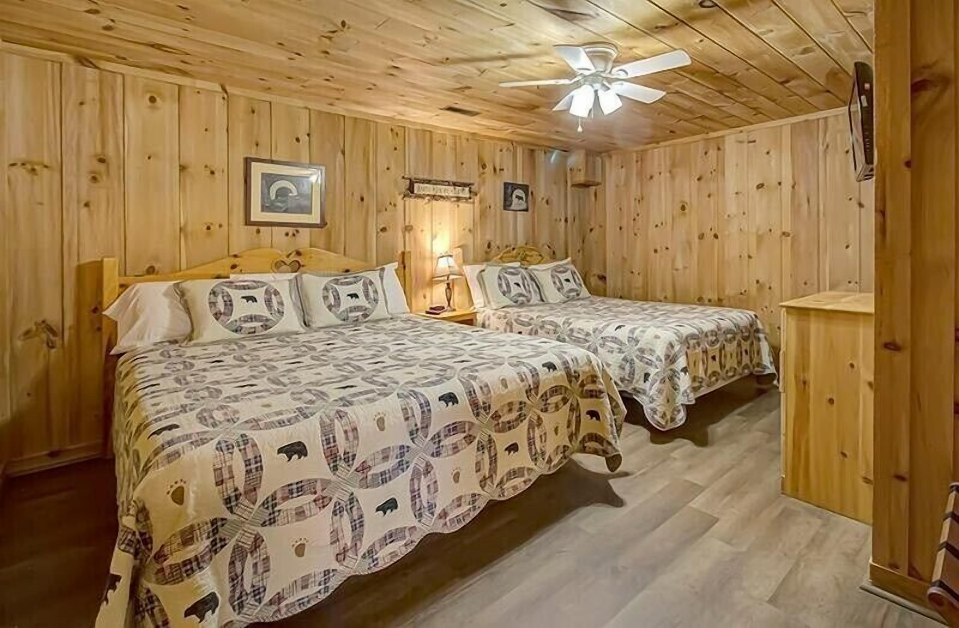 Una habitación tipo cabaña con dos camas, paredes de madera, un ventilador de techo y una pequeña mesa.
