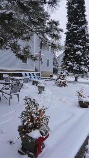 A snow-covered outdoor area with a house, patio furniture, and Christmas trees.