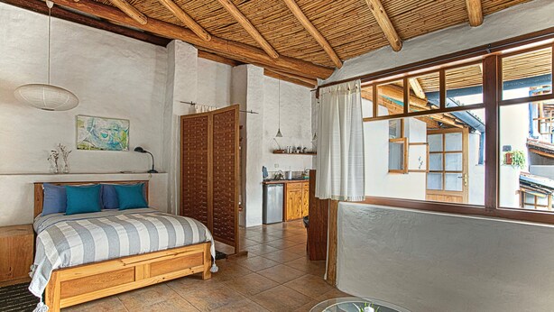 A bedroom with a wooden bed, a small table, and a window with curtains. - Studio Mariposa with beautiful gardens in Historical Quito (Quito)