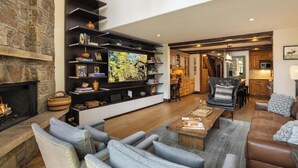A spacious living area with a fireplace, comfortable seating, a kitchen in the background, and a large TV on a wall.