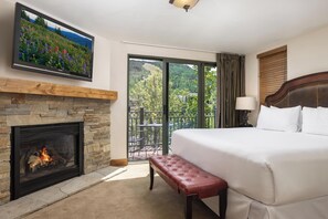 Ein Hotelzimmer mit Kamin aus Stein, einem großen Bett, einer Bank, einem an der Wand montierten Fernseher und einem Balkon mit Blick auf Grünflächen und Berge.