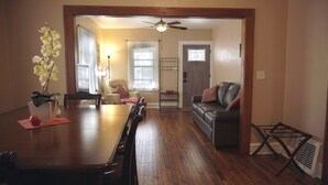 A dining area with a wooden table, chairs, and a vase of flowers. There is a living room with a sofa and a ceiling fan. - CORNER OF CALM IN THE ♥ OF CEDAR RAPIDS! (Cedar Rapids)