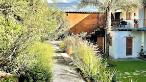A garden pathway leading to a white building with a balcony, surrounded by lush greenery and a mountain backdrop. - Cosy maisonette in a beautiful garden - historic ambience and modern comfort (Quito)