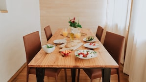 A wooden dining table with chairs, plates of food, a bottle of olive oil, glasses of orange juice, and a vase with flowers. - Cosy maisonette in a beautiful garden - historic ambience and modern comfort (Quito)
