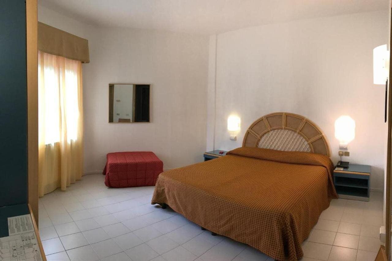 Une chambre à coucher avec un lit, une table de chevet, un pouf rouge, un miroir et une fenêtre avec des rideaux.