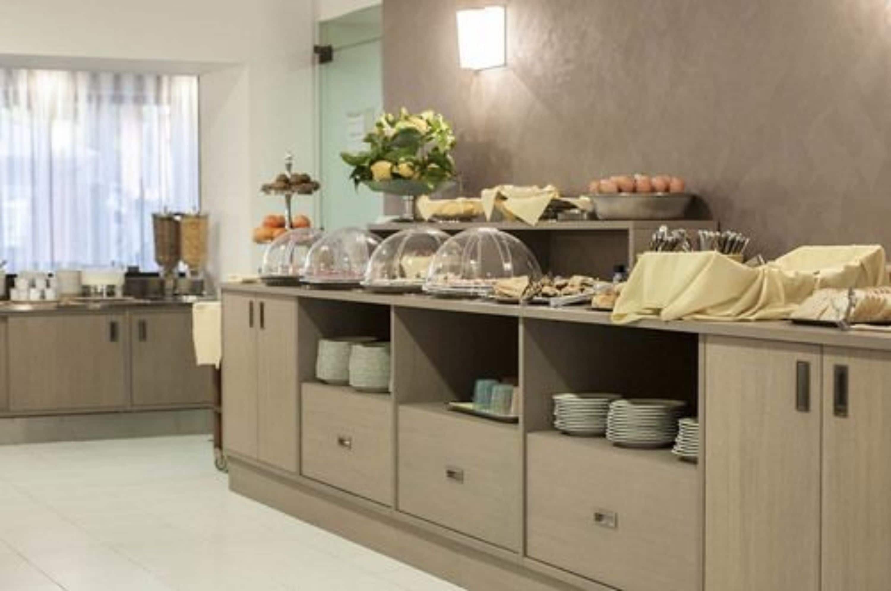 Habitación de hotel con kitchenette, zona de comedor con mesas de buffet y un amplio ventanal con cortinas.