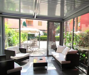 Een moderne leefruimte, zowel binnen als buiten, met een glazen wand, een zithoek, een salontafel en een eethoek met stoelen en een parasol.