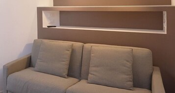Un sofá color beige con dos cojines frente a una pared con un estante encima.