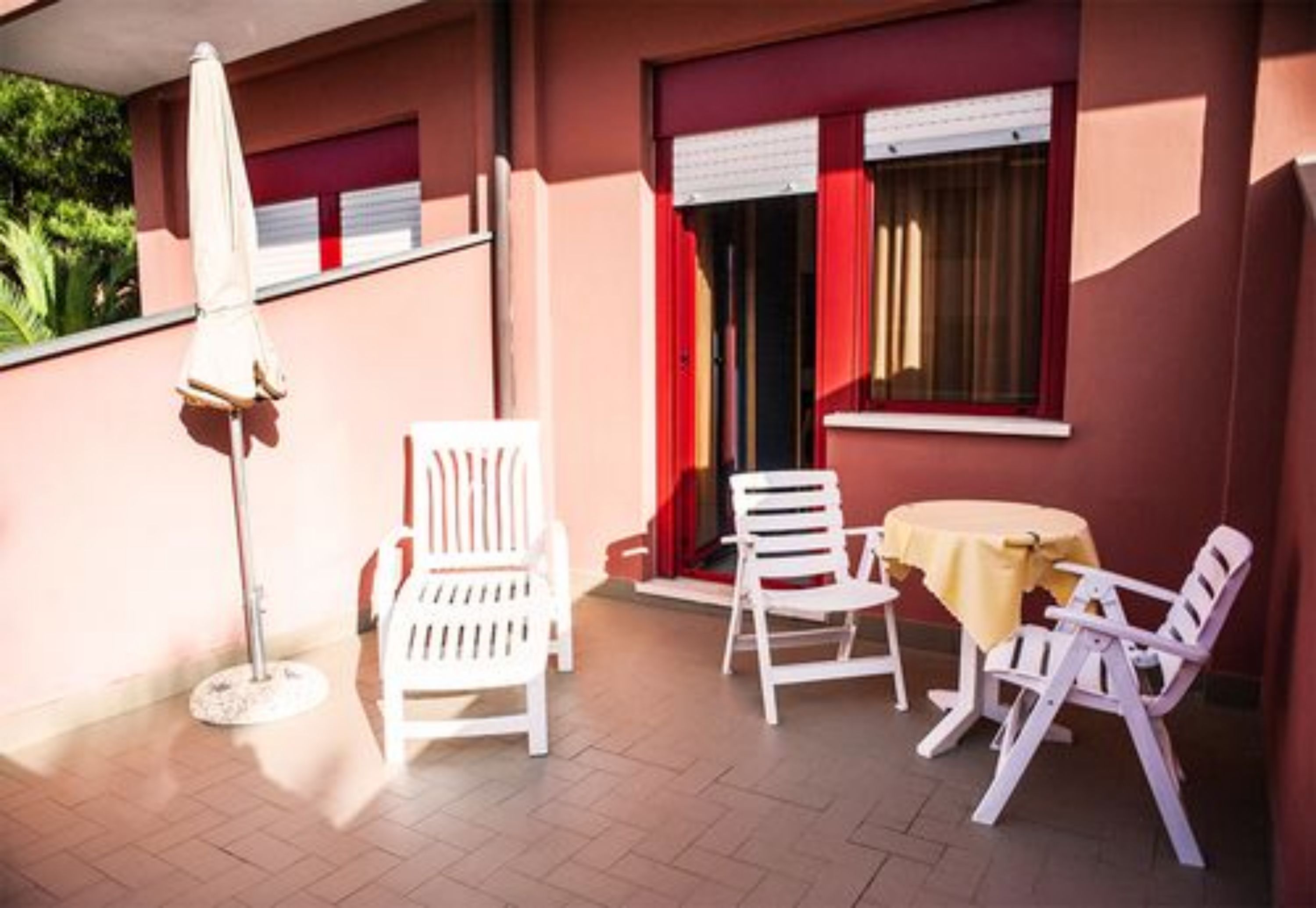 Un patio con sillas y mesa de plástico blanco, un paraguas blanco y una ventana con cortinas rojas.
