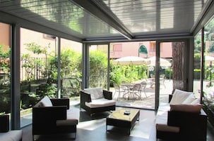 A conservatory with a glass ceiling, furnished with sofas and a coffee table, overlooking a patio area with outdoor seating and greenery.