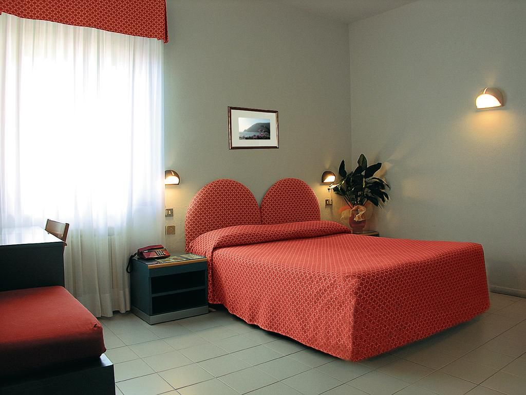 A bedroom with a bed, a desk, a chair, a nightstand, a telephone, a framed picture on the wall, and two wall-mounted lights.