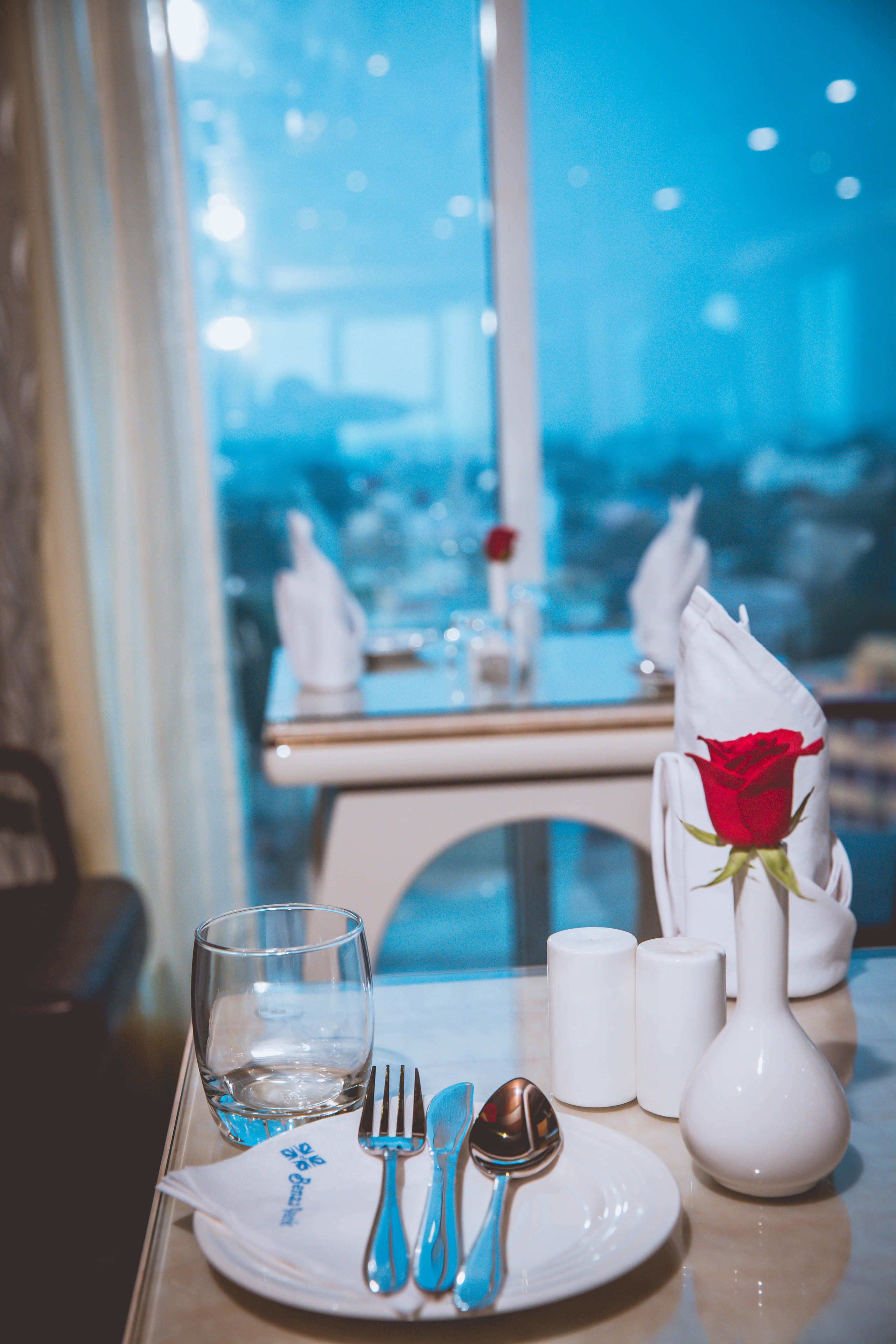 A dining table set with a plate, cutlery, a glass, and a vase with a rose.