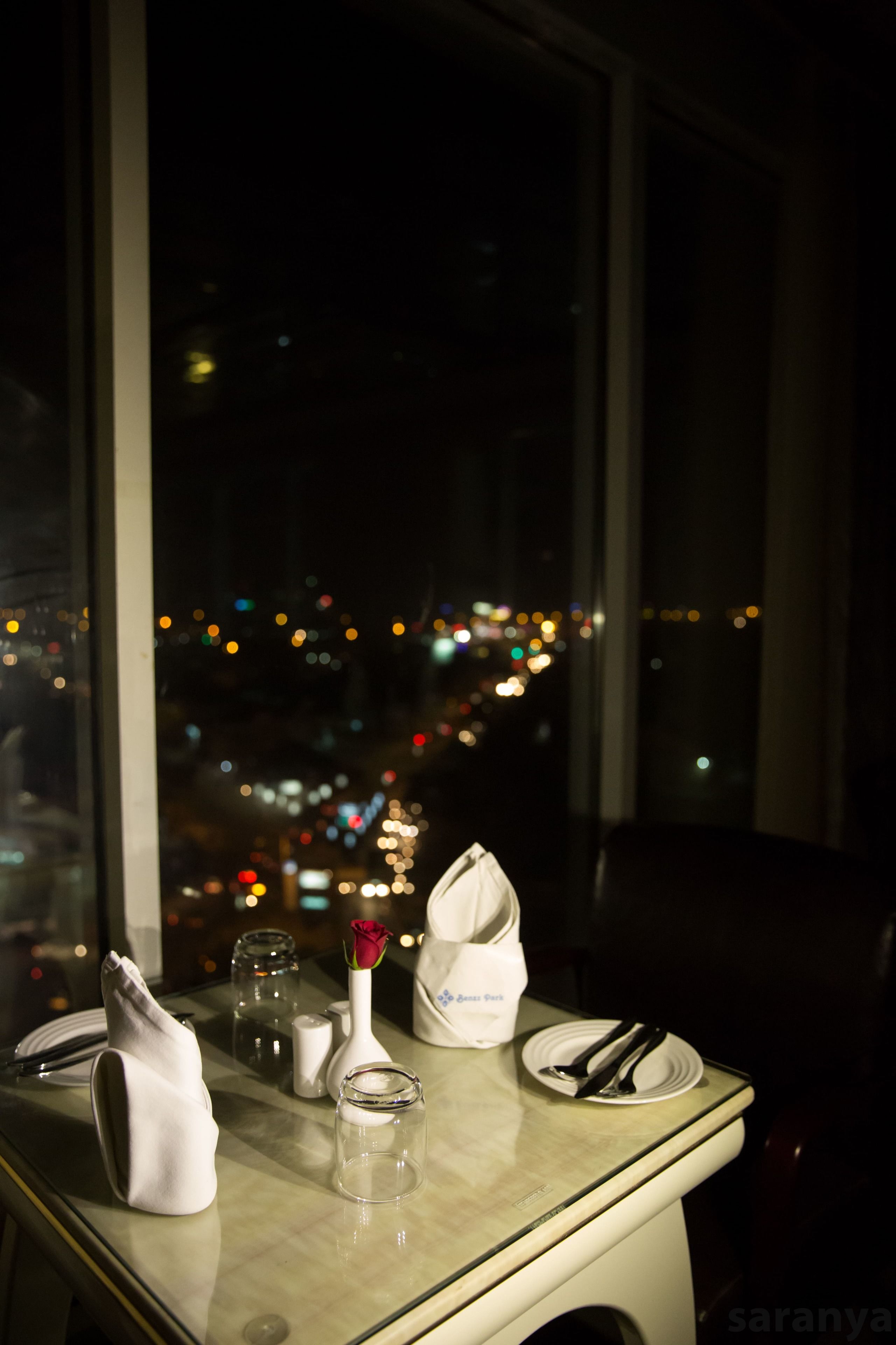 A table set with a white napkin, a glass, a vase with a single rose, and a plate with cutlery, viewed through a window overlooking a cityscape at night.
