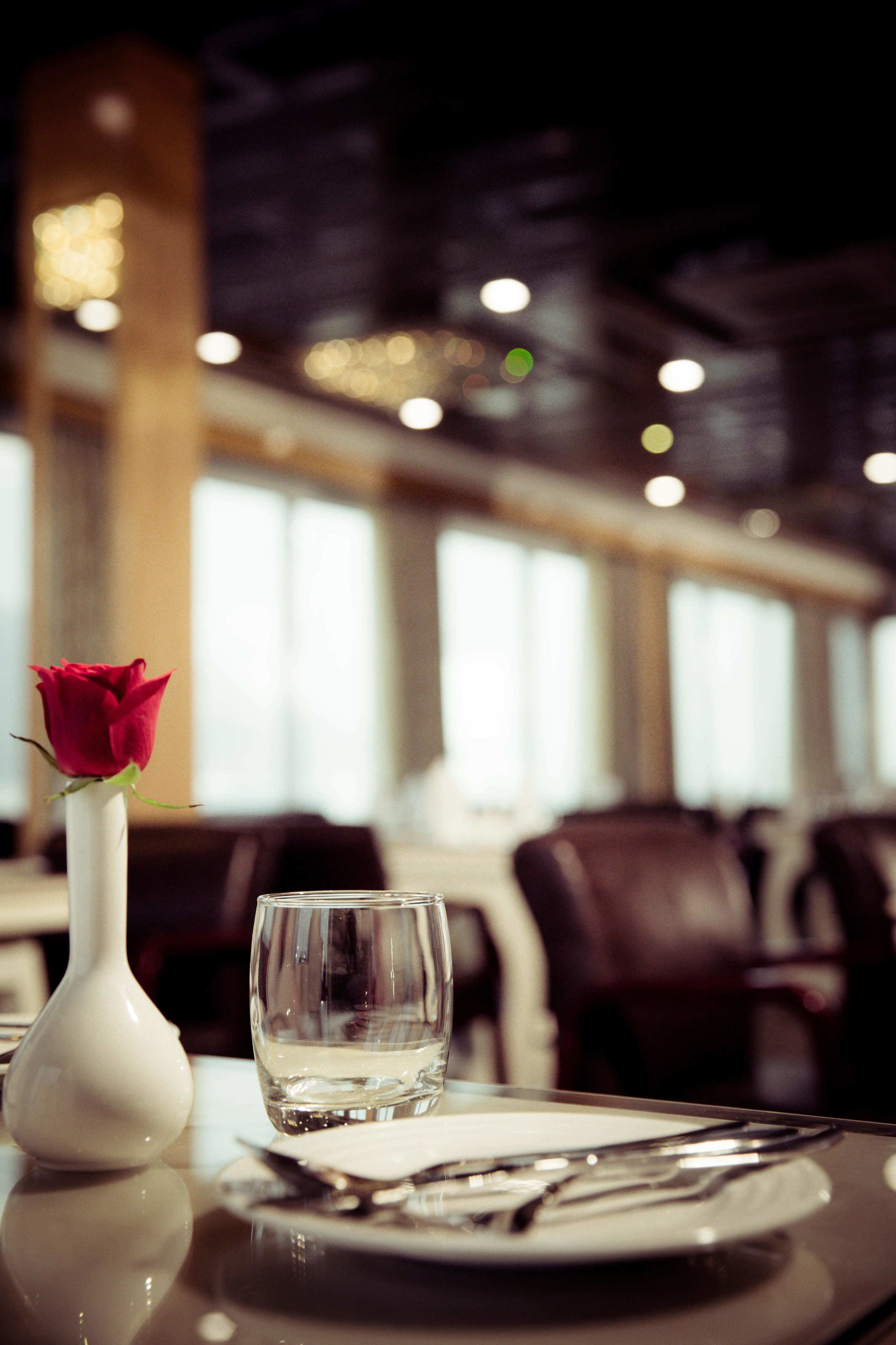 A dining table with a white vase containing a red rose, a clear glass, and neatly arranged cutlery.