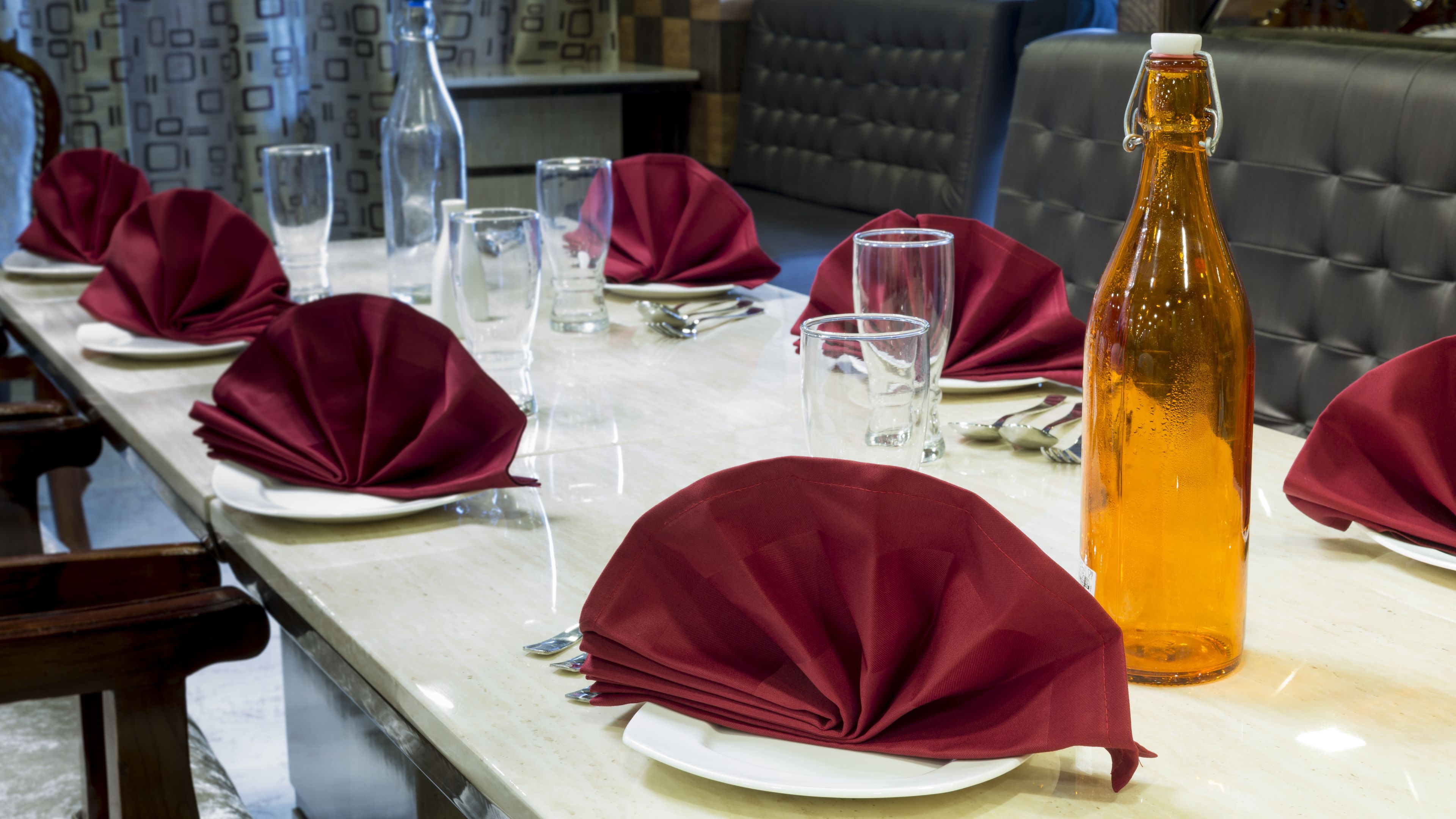 A dining table set with red napkins, clear glasses, and a bottle of amber liquid.