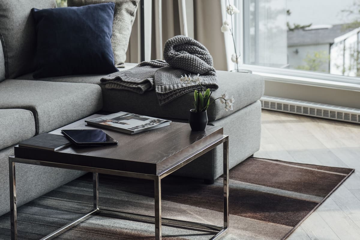 Un salon moderne avec un canapé gris, une table basse et un lampadaire. Une grande fenêtre offre une vue sur un lac et la verdure environnante.