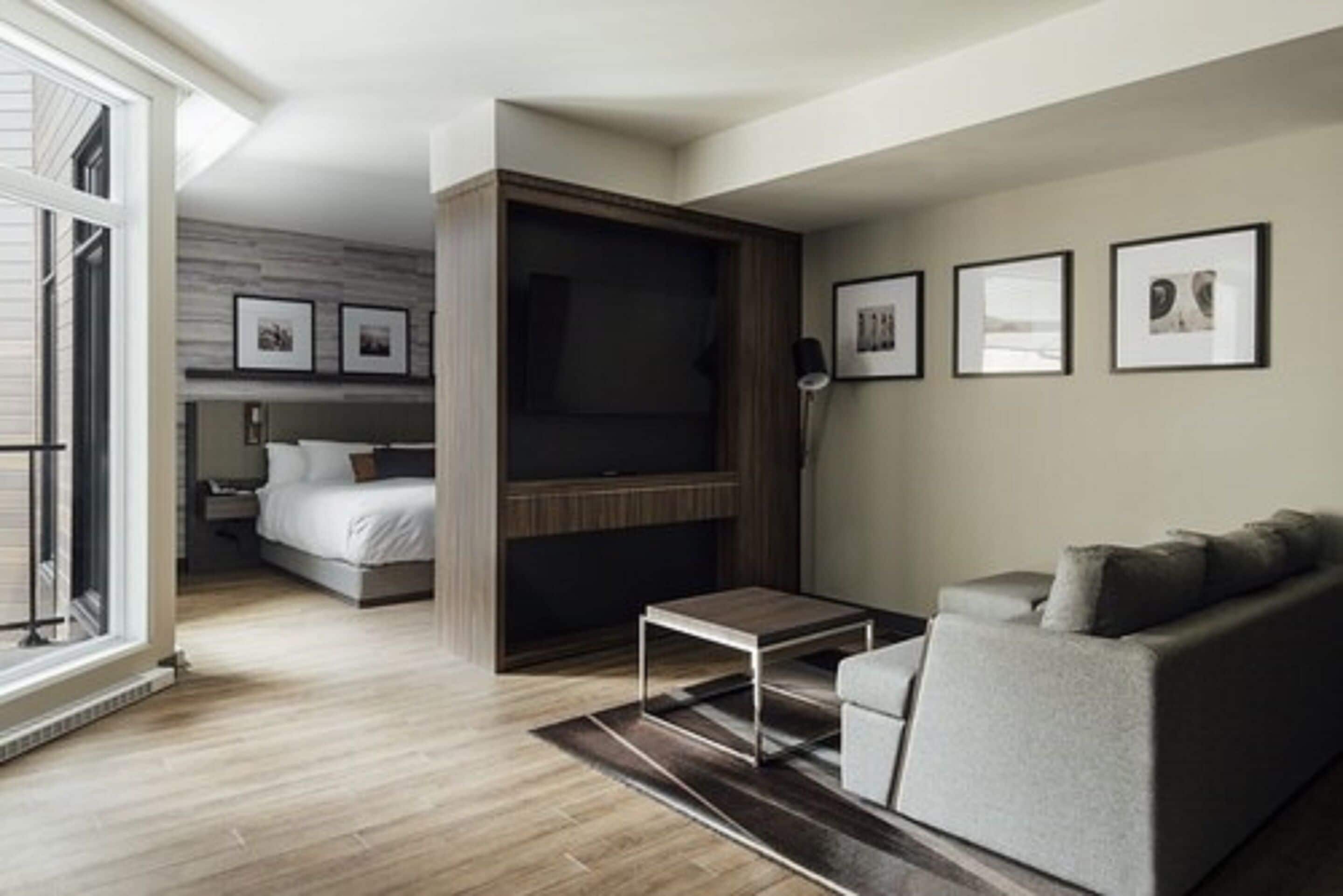 A modern living room with a sofa, a coffee table, and a TV mounted on a wooden wall. There is a bed with a headboard in the background and a window with blinds.