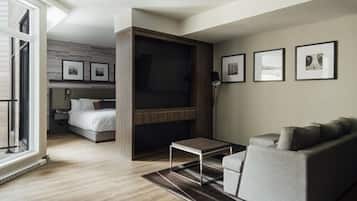 A modern living room with a sofa, a coffee table, and a TV mounted on a wooden wall. There is a bed with a headboard in the background and a window with blinds.