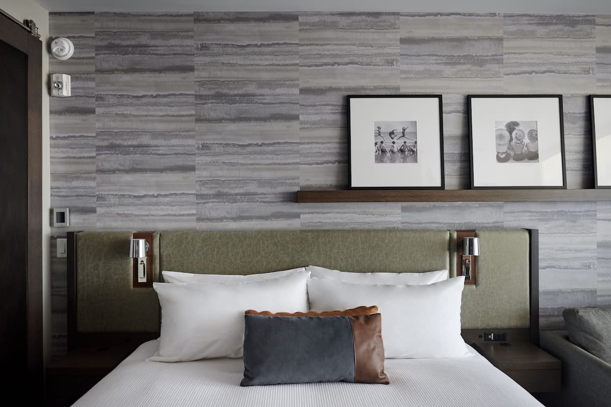Une chambre d’hôtel avec un lit, des oreillers, une tête de lit, une table de chevet, une lampe et des tableaux encadrés au mur.