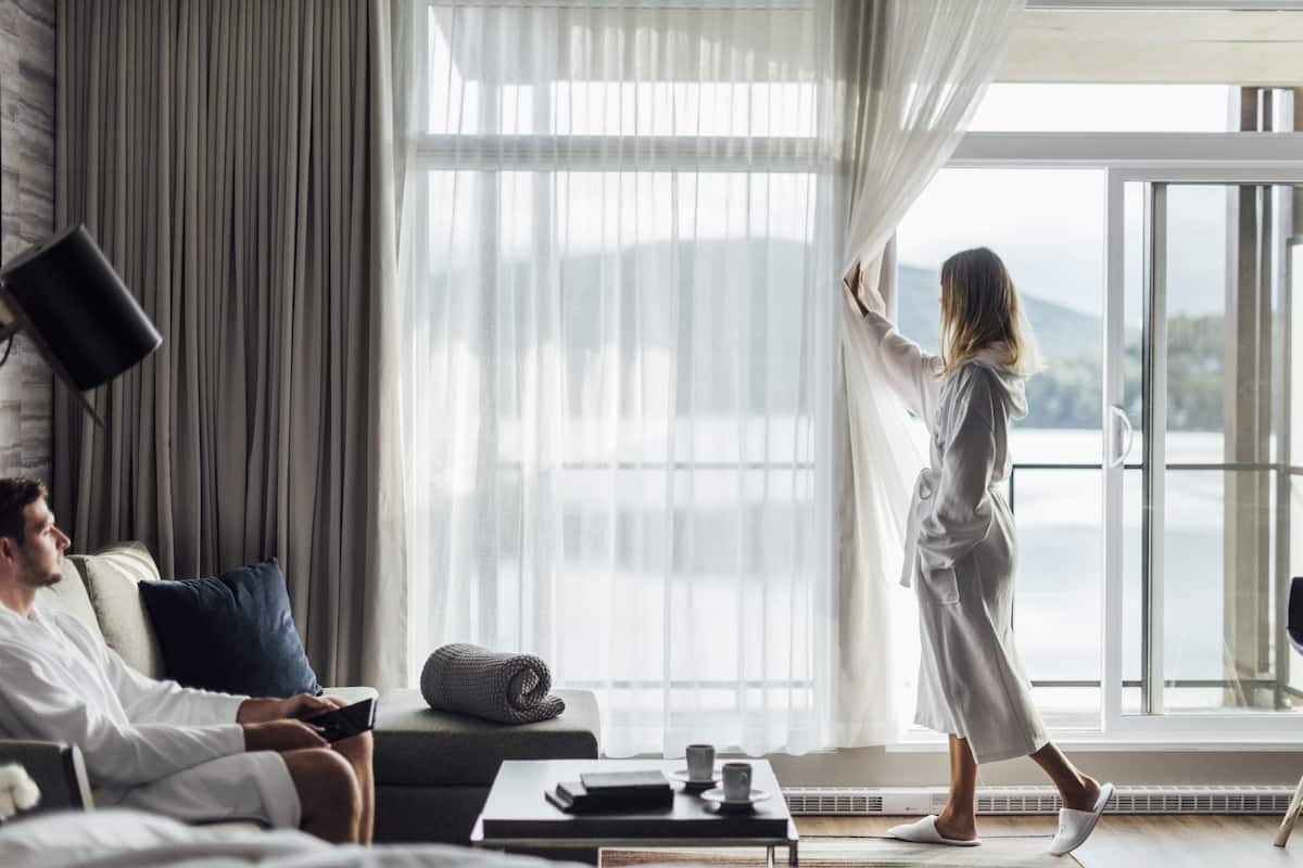 Une chambre d’hôtel moderne avec une grande fenêtre, un canapé, une table basse et une personne portant une robe de chambre.
