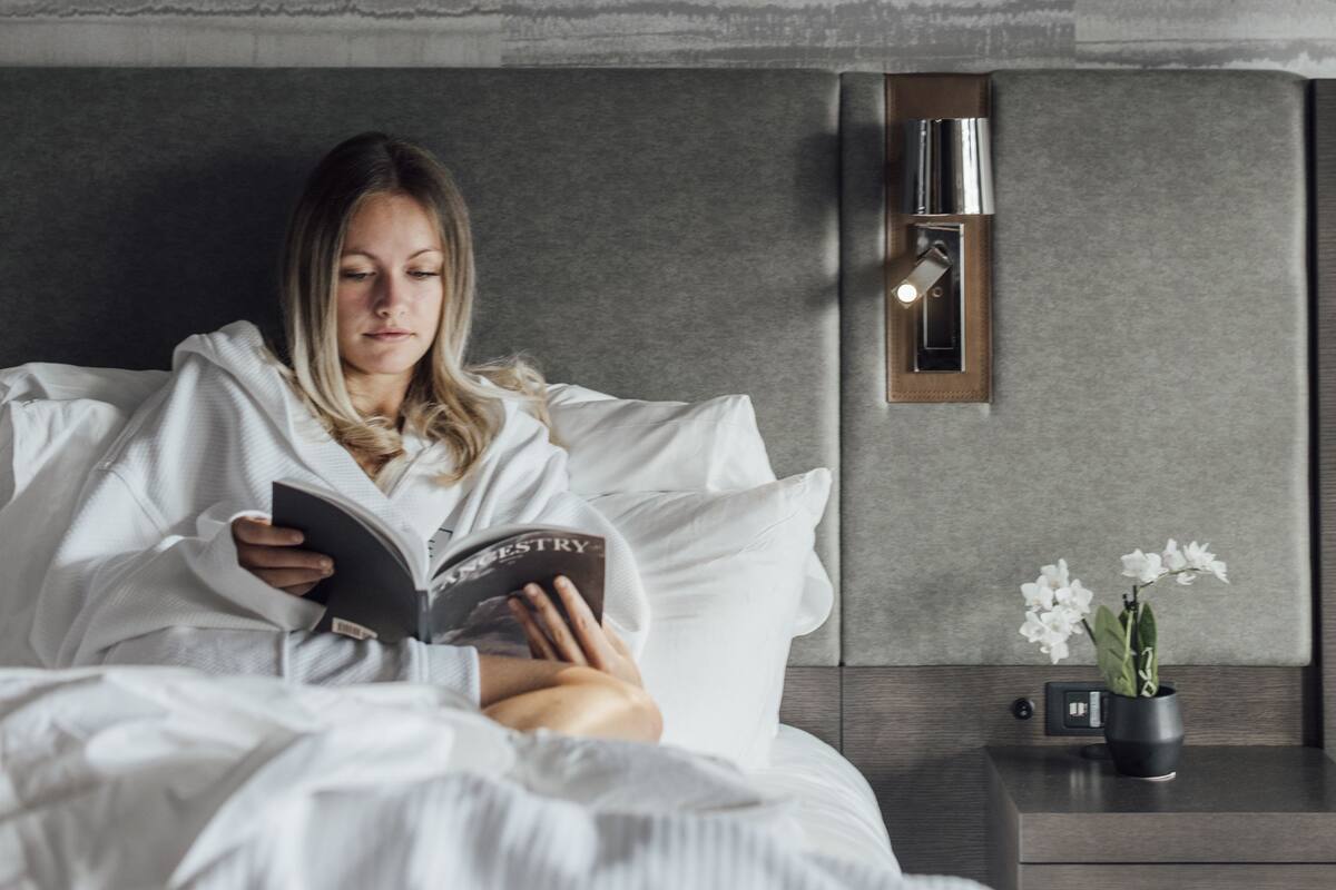 Une personne est assise, lisant un livre, dans une chambre moderne. On y voit un mur gris à texture, un tableau encadré au-dessus du lit, une table de chevet avec une lampe et une petite plante en pot.