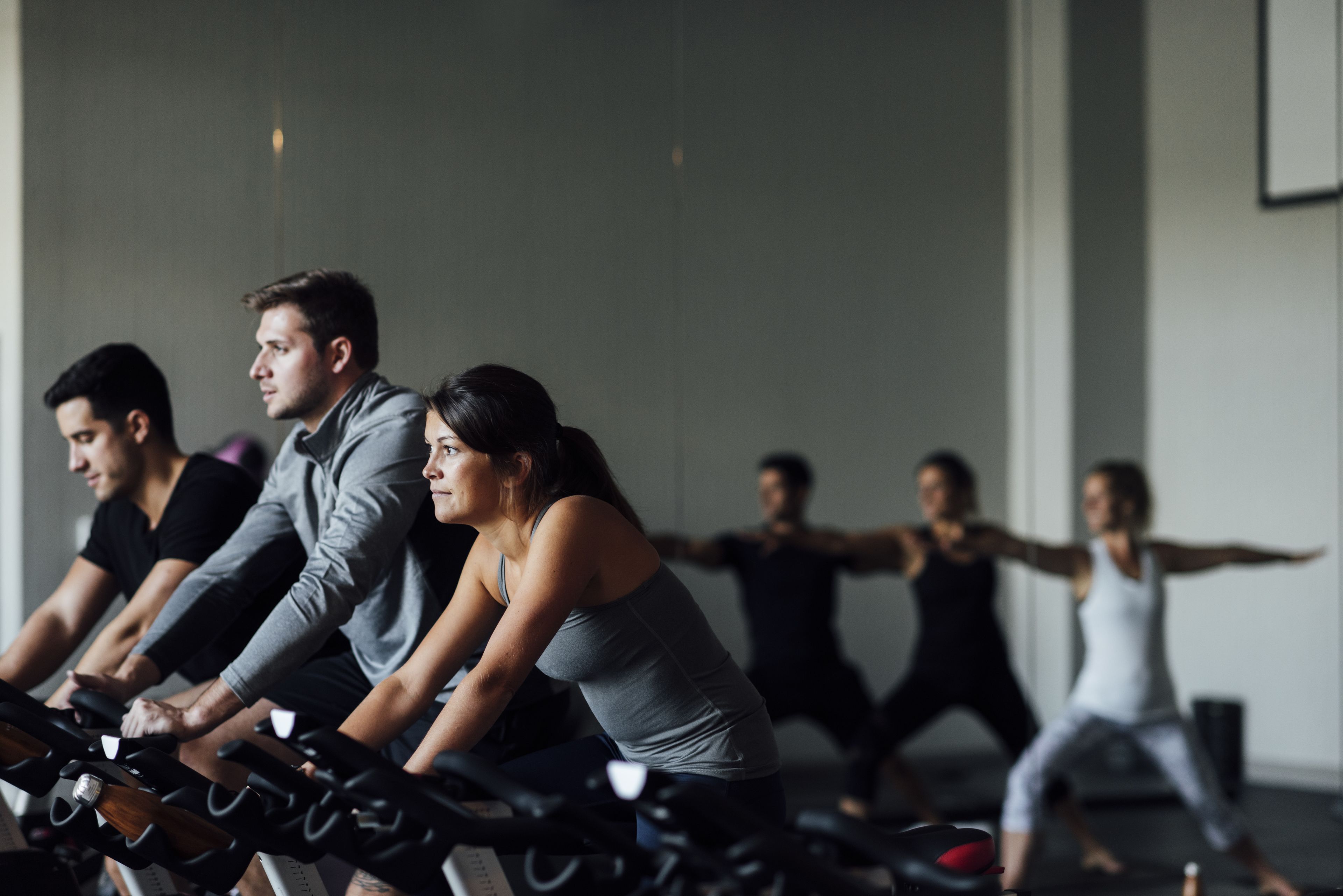 A group of people exercising on stationary bikes in a gym with a plain wall and a door in the background.