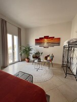 Una stanza ben illuminata con un'ampia finestra, un letto, una zona salotto con tavolo e sedie, e un'opera d'arte appesa al muro.