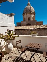 Una terrazza con un muro bianco, una pianta di cactus, una panchina di legno, un tavolo e vista su un edificio storico con una cupola.