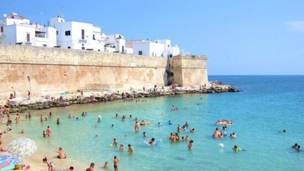 A beach with people swimming and sunbathing, white buildings in the background, and a clear blue sky. - Nenetta Rooms (Monopoli)