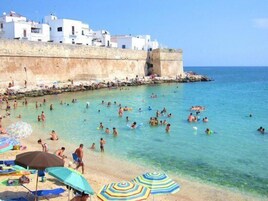 Una spiaggia con persone che nuotano e prendono il sole, edifici bianchi sullo sfondo e un cielo azzurro limpido.