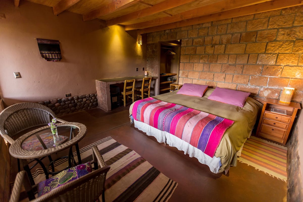 A cozy room with a bed, a small table with chairs, a wooden bench, and a stone wall.