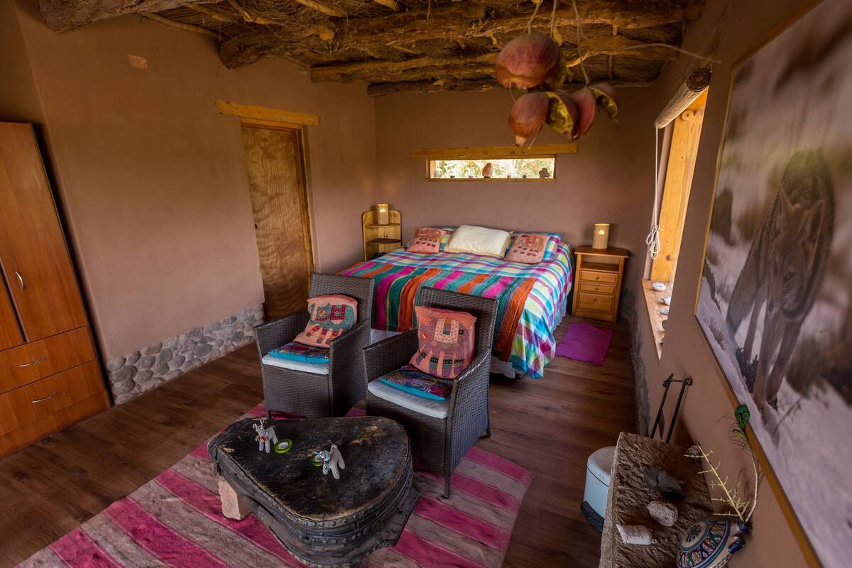 A rustic bedroom with a bed, two wicker chairs, a small table, a wooden wardrobe, and a stone wall.