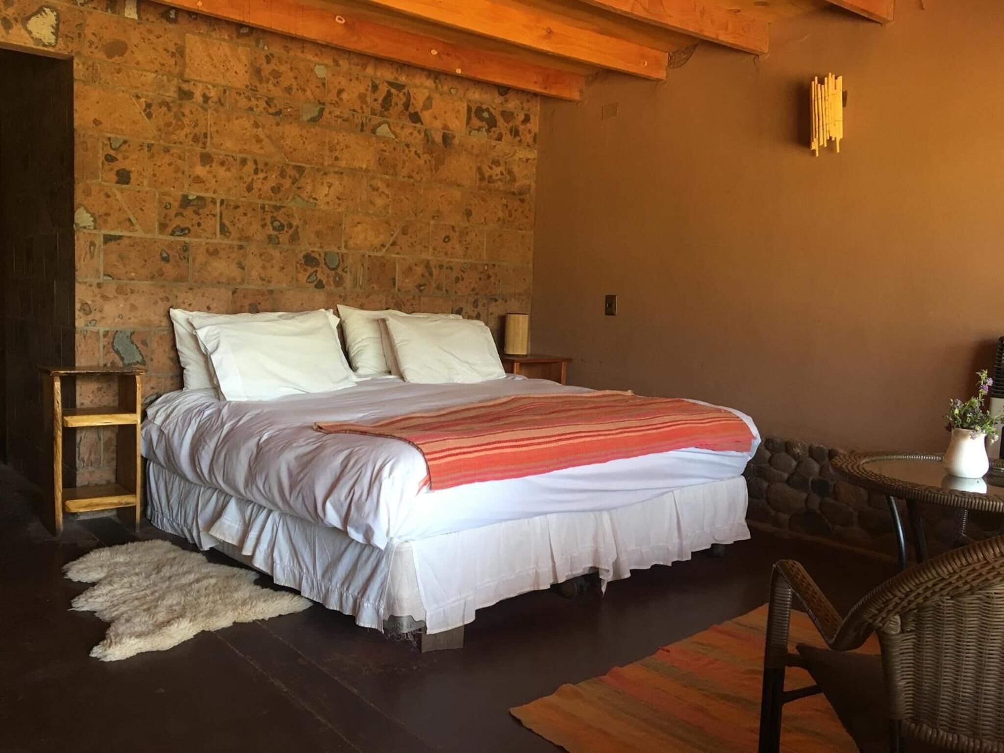 Un dormitorio con pared de piedra, cama con ropa blanca, mesita de noche de madera, una pequeña mesa con una planta y una silla de mimbre.