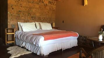 Un dormitorio con pared de piedra, cama con ropa blanca, mesita de noche de madera, una pequeña mesa con una planta y una silla de mimbre.