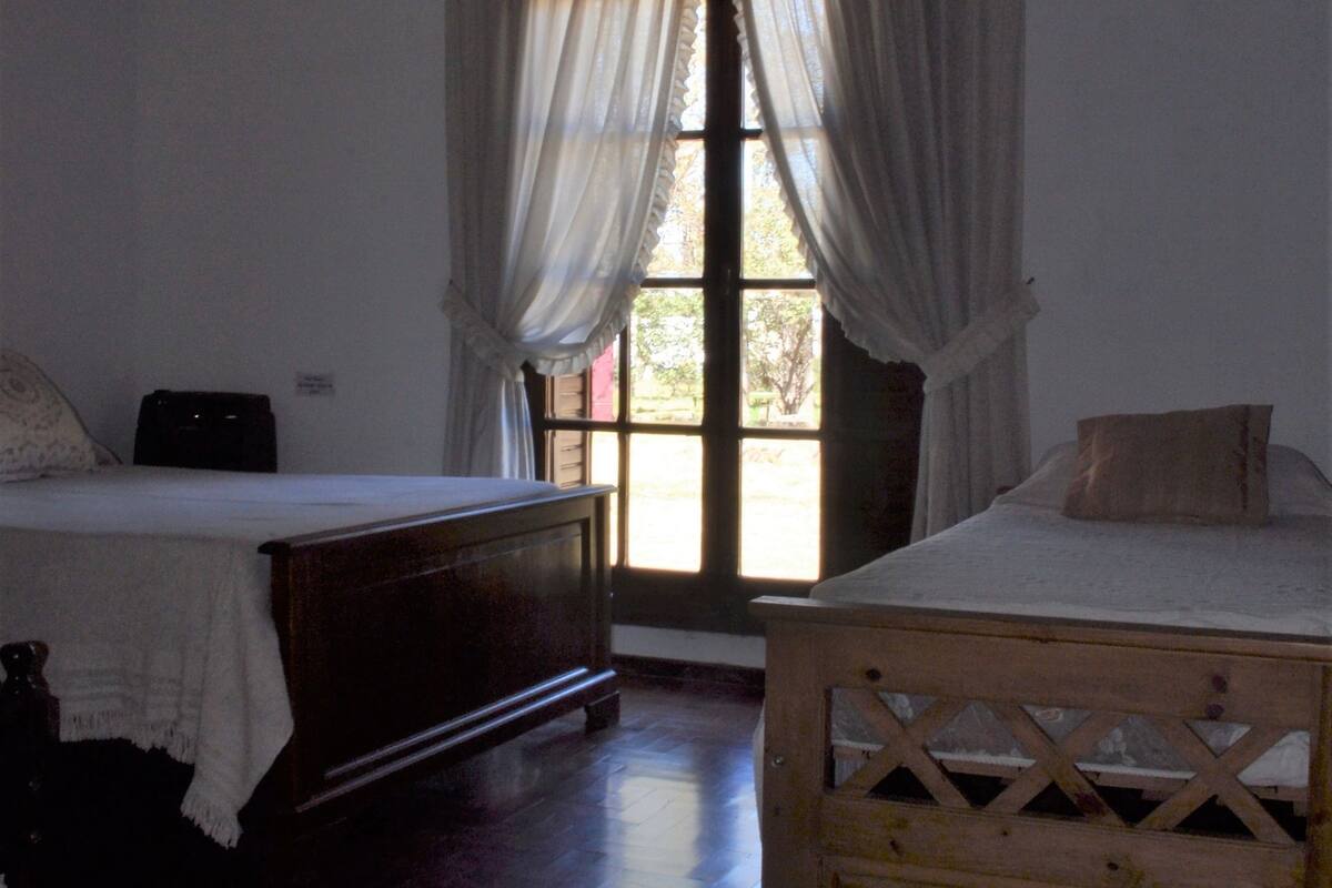Ein Zimmer mit zwei Holzbetten, einem Deckenventilator und einem großen Fenster mit Vorhängen.