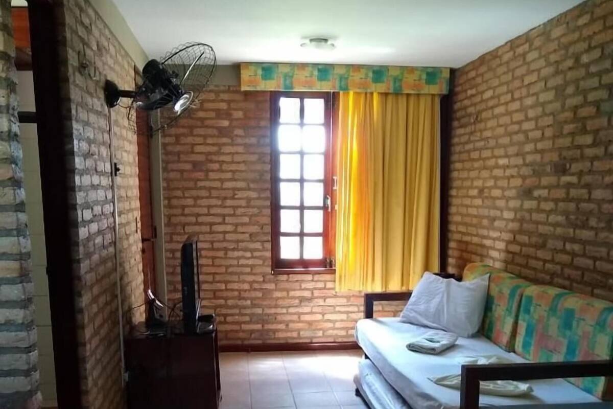 A room with a brick wall, a bed with a colorful bedspread, a sofa with a white pillow, a window with yellow curtains, and a standing fan.