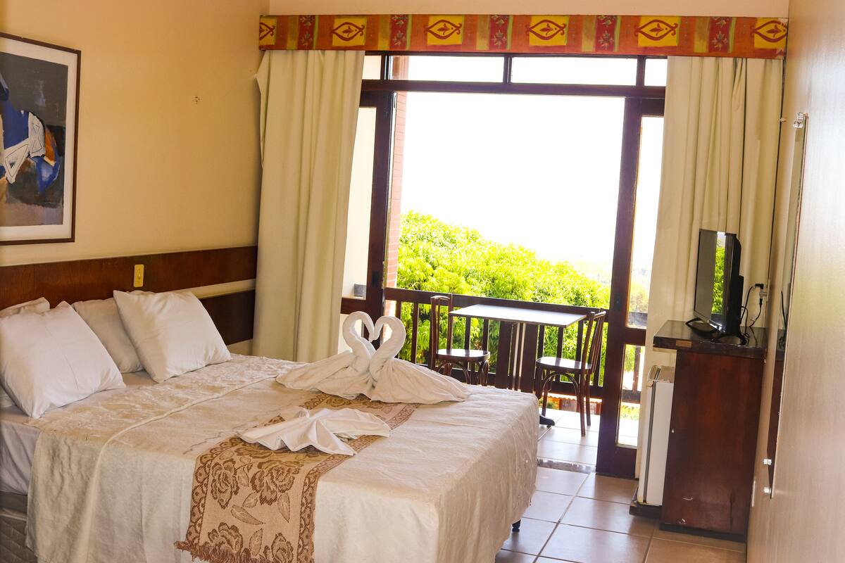 A hotel room with a bed, a desk, a TV, and a balcony with a view of greenery.