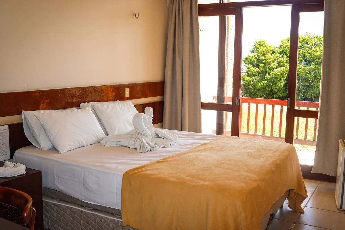 A hotel room with a bed, a wooden headboard, a window with a view of greenery, and a balcony with a wooden railing.