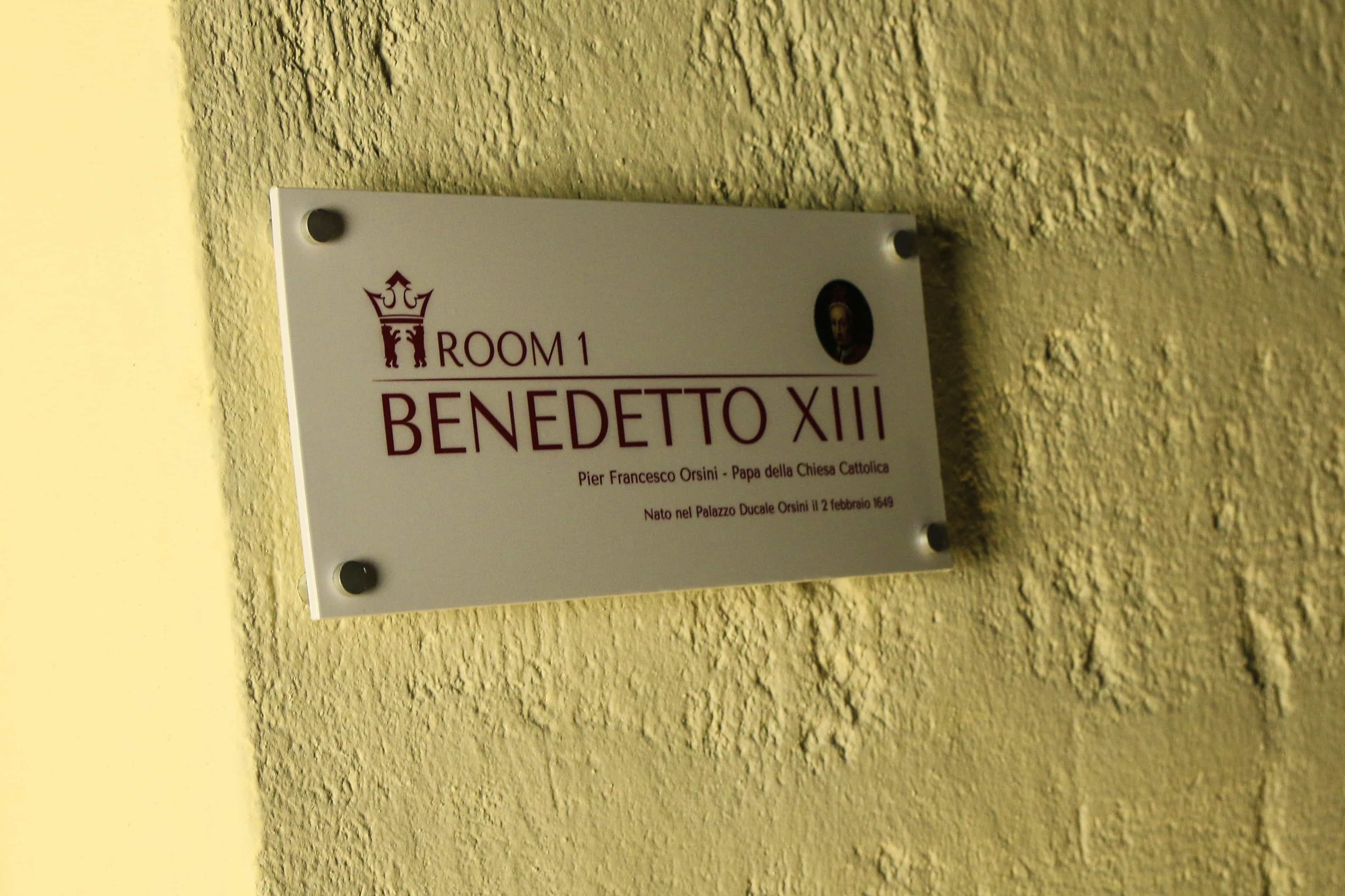 A plaque on a wall with the text "ROOM 1 BENEDETTO XIII Pier Francesco Orsini - Papa della Chiesa Cattolica Nato nel Palazzo Ducale Orsini il 2 febbraio 1451".