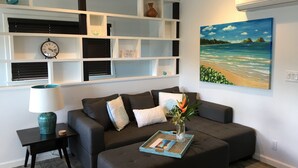 A living room with a dark grey sofa, a small side table with a lamp, a wall-mounted shelf with decorative items, and a painting of a beach scene.