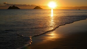 A beach scene with the sun setting over the ocean, waves gently lapping at the shore, and distant islands in the background.