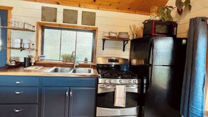 A compact kitchen with a stove, oven, and refrigerator, wooden cabinets, and a window with blinds.